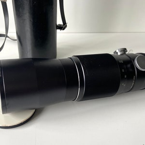 Puede incluir: Un objetivo teleobjetivo negro con un anillo plateado y una tapa de lente negra. El objetivo est&aacute; sobre una superficie blanca.