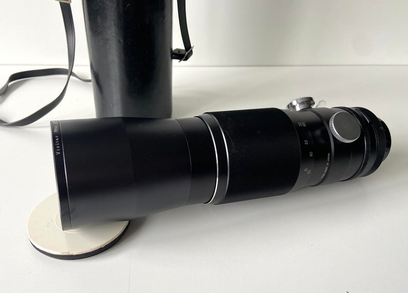 Puede incluir: Un objetivo teleobjetivo negro con un anillo plateado y una tapa de lente negra. El objetivo est&aacute; sobre una superficie blanca.