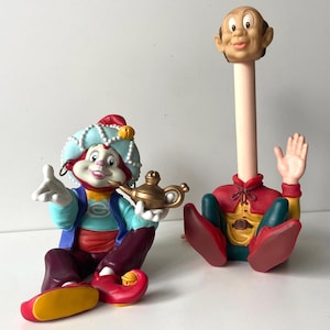 May include: Two whimsical figurines. One is a genie-like character with a blue and red turban, holding a golden lamp. The other is a cartoon figure with an elongated neck, wearing a red and yellow outfit, with a raised hand.