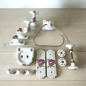 May include: A collection of white ceramic bathroom accessories featuring a pink rose floral design. The set includes towel rings, soap dishes, toothbrush holders, and door knobs. The accessories are arranged on a light wood surface.