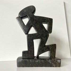 May include: A dark gray stone sculpture of a person kneeling, with a hand on the head. The figure is stylized with geometric shapes and is mounted on a rectangular base. The sculpture has a textured surface.