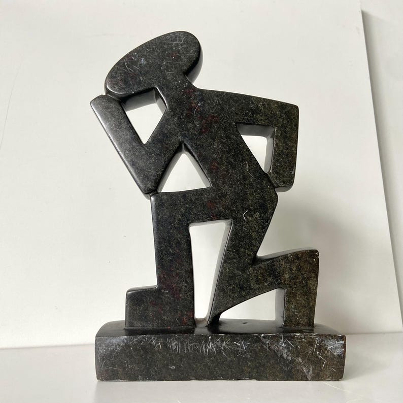 May include: A dark gray stone sculpture of a person kneeling, with a hand on the head. The figure is stylized with geometric shapes and is mounted on a rectangular base. The sculpture has a textured surface.