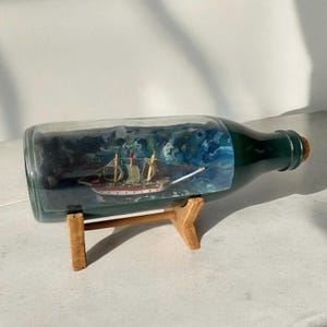 Puede incluir: Una botella de vidrio con un exterior verde y un soporte de madera. Dentro de la botella hay un barco en miniatura navegando en un mar pintado de azul y blanco.