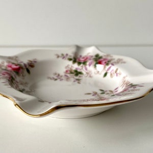 May include: A white porcelain trinket dish with a scalloped edge and a floral design in shades of pink, purple, and green. The dish has a gold trim around the edge.