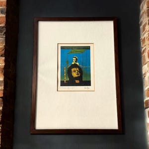 May include: Framed art print featuring a surrealist image. The artwork depicts a figure with a dark, curly hairstyle and a second figure above, set against a blue and green background. The print is framed in a dark wood frame.