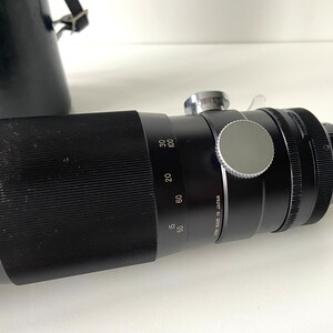 Puede incluir: Un objetivo teleobjetivo negro con un anillo plateado y una tapa de lente plateada. El objetivo tiene marcas para 50, 60, 80, 100 mm y "Lens Made in Japan".