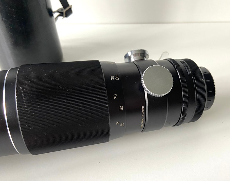 Puede incluir: Un objetivo teleobjetivo negro con un anillo plateado y una tapa de lente plateada. El objetivo tiene marcas para 50, 60, 80, 100 mm y "Lens Made in Japan".