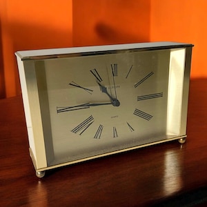May include: A rectangular quartz clock with a gold-colored face and black Roman numerals. The clock has a white and gold frame with small, round feet. The word "EUROPA" is printed above the clock hands.