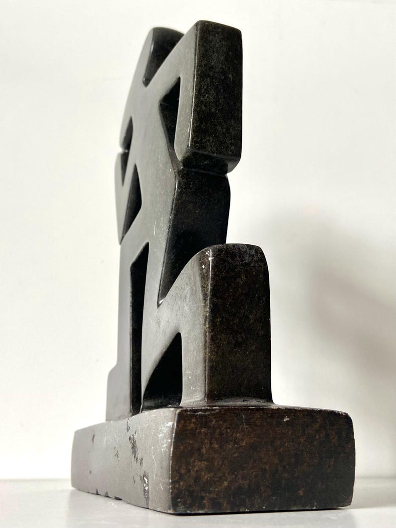 May include: A dark, abstract sculpture with a geometric design. The sculpture is made of a dark, possibly stone-like material and features a series of interconnected shapes. It stands on a rectangular base, and the overall form is angular and modern.