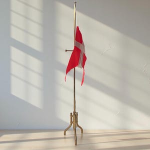 May include: A brass flag stand with a red and white flag. The flag has a white Scandinavian cross on a red background. The stand has a three-legged base and a tall pole. The flag is slightly tattered.