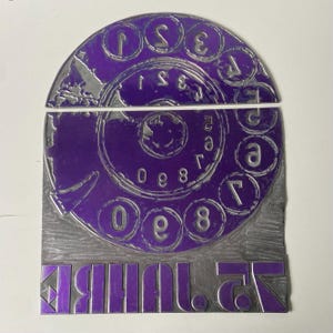 Puede incluir: Un dial de teléfono rotatorio morado y plateado con la palabra "DIALER" impresa al revés en la parte inferior. El dial presenta números y letras, con un diseño circular.