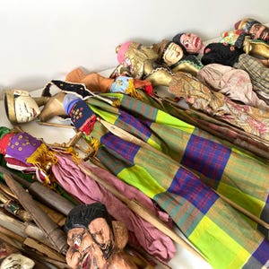 May include: A collection of multiple rod puppets featuring hand-painted wooden heads with distinct facial expressions, adorned in colorful fabric garments including plaid patterns, solid pinks, and gold accents, each attached to thin wooden control rods.