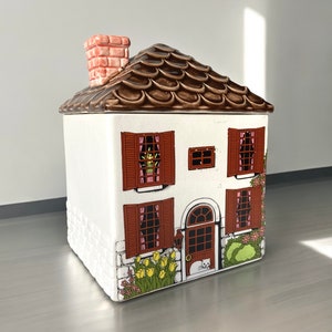 Puede incluir: Una caja de galletas de cerámica blanca con forma de casa con un techo marrón, contraventanas marrones y un gato sentado en el porche. La casa tiene una chimenea y un jardín de flores.