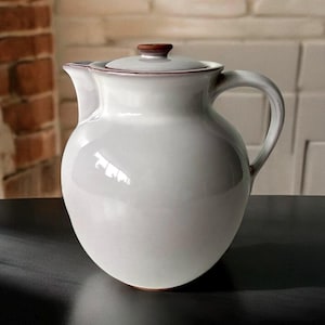 May include: A white ceramic teapot with a brown lid and a rounded body. The teapot has a handle on the side and a spout on the front.