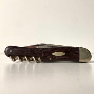 以下が含まれることがあります： ダークブラウンの木目調ハンドルを備えた折りたたみ式ポケットナイフ。片端には金属製のボルスターがあり、側面には銀色のコルク抜きが見えます。ハンドルの中央には、小さな楕円形の金属製インレイが埋め込まれています。