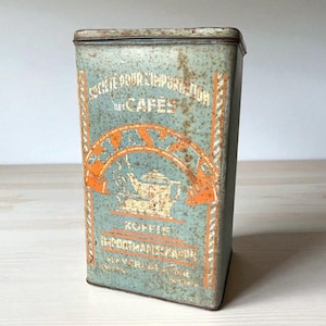 Op de afbeelding: Vintage rechthoekige metalen koffieblik met een vervaagd teal en oranje ontwerp. Het blik bevat tekst in het Frans en Nederlands, waaronder "SOCIETE POUR L'IMPORTATION DES CAFES" en "KOFFIE". Het ontwerp bevat een koffiepot.