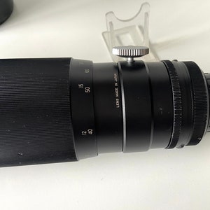 Puede incluir: Un objetivo de c&aacute;mara negro con un anillo de enfoque plateado y una tapa de lente negra. El objetivo tiene marcas para diferentes distancias focales, incluyendo 12, 40, 50 y 60. El objetivo est&aacute; etiquetado como "Lens Made in Japan".