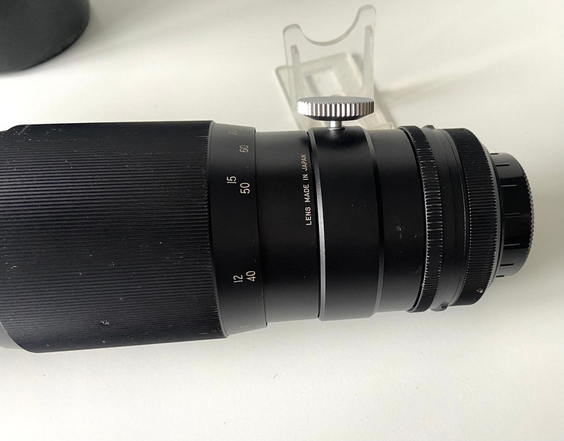 Puede incluir: Un objetivo de c&aacute;mara negro con un anillo de enfoque plateado y una tapa de lente negra. El objetivo tiene marcas para diferentes distancias focales, incluyendo 12, 40, 50 y 60. El objetivo est&aacute; etiquetado como "Lens Made in Japan".