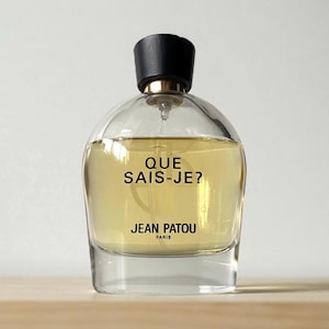 Op de afbeelding: Een heldere glazen parfumfles met een zwarte dop en gouden accenten. De fles bevat een licht amberkleurige vloeistof en heeft de tekst "QUE SAIS-JE?" en "JEAN PATOU PARIS". De fles staat op een lichtbruin oppervlak.
