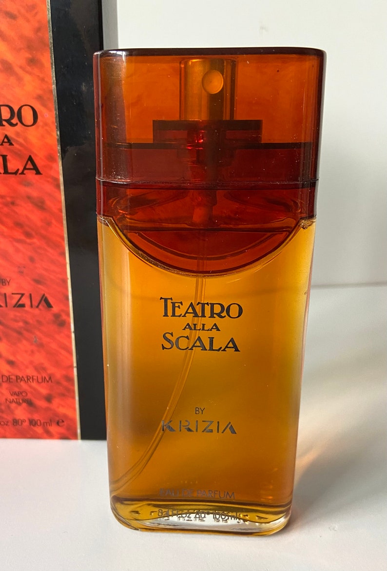 Pu&ograve; includere: Un flacone di profumo Teatro Alla Scala di Krizia in vetro marrone. Il flacone ha un'etichetta dorata con testo nero.