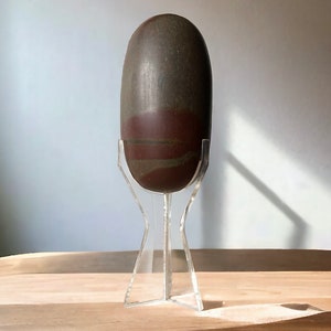 以下が含まれることがあります： 滑らかで研磨された表面を持つ、大きな楕円形の石。石は主に濃い茶色で、赤みがかった茶色と灰色の帯があります。透明なアクリルスタンドに展示されています。装飾品です。