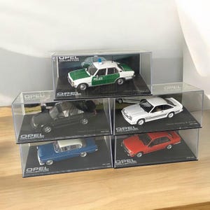 Op de afbeelding: Vijf miniatuur automodellen in vitrinekasten. De auto's zijn allemaal Opel-modellen, en elke vitrine heeft het Opel Collection-logo. De auto's zijn een witte Opel Manta, een rode Opel Kadett, een blauwe Opel Rekord, een zwarte Opel Ascona en een groene Opel Rekord politieauto.