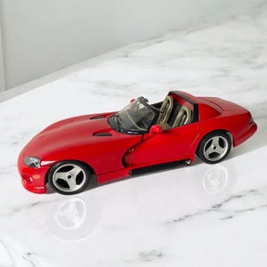 Puede incluir: Un modelo de coche rojo de juguete de un Dodge Viper descapotable. El coche tiene detalles negros y asientos interiores de color beige. El coche está sobre una superficie de mármol blanco.