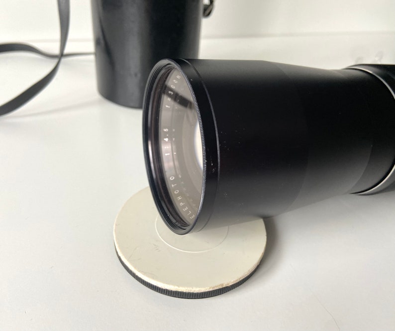 Puede incluir: Un objetivo de c&aacute;mara negro con un anillo plateado y marcas alrededor del borde. El objetivo est&aacute; sobre una superficie circular blanca.