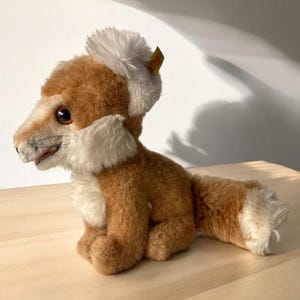 May include: A soft toy fox with brown and white fur, including a bushy tail and ear accents. The fox has a detailed face with dark eyes and a stitched nose. The toy is sitting on a light-coloured wooden surface.