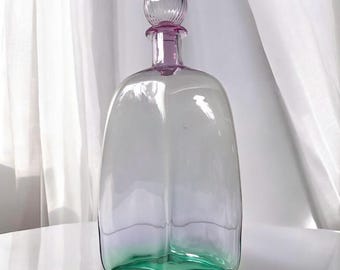 May include: A clear glass bottle with a pink stopper and a green base. The bottle is square shaped and has a textured surface.