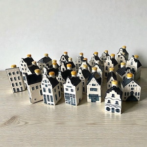 May include: A collection of miniature ceramic house-shaped bottles, each featuring a cream-coloured facade with dark blue architectural details and a small yellow cap on the roof. These decorative items vary in design, representing different building styles.
