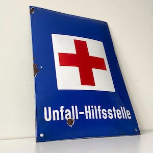 Puede incluir: Letrero metálico azul vintage con un cuadrado blanco y una cruz roja. El texto "Unfall-Hilfsstelle" es blanco. El letrero muestra signos de desgaste y óxido.