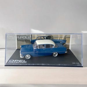 Könnte beinhalten: Ein blaues und weißes Opel-Automodell von 1957-1960 in einer transparenten Vitrine. Die Vitrine hat eine schwarze Basis mit dem aufgedruckten Text "Opel Collection".