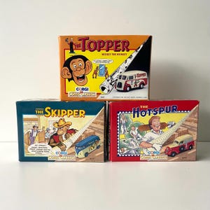 Puede incluir: Tres cajas de coches de juguete vintage con personajes de cómics británicos. Las cajas son rojas, naranjas y azules con ilustraciones de los personajes y el texto "The Topper", "The Skipper" y "The Hotspur".