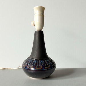 Könnte beinhalten: Eine schwarze Keramik-Tischlampe mit cremefarbener Fassung und einem dekorativen Muster um den Sockel. Die Lampe hat eine tropfenförmige Form und einen langen, schlanken Hals. Der Sockel weist ein sich wiederholendes Muster in Blau-, Braun- und Rottönen auf.