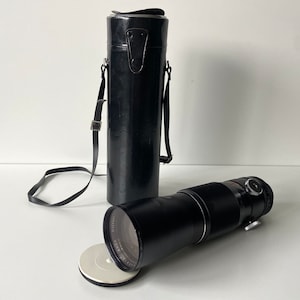 Puede incluir: Un objetivo de c&aacute;mara negro con una funda de transporte de cuero negro. El objetivo est&aacute; sobre una superficie blanca con una tapa circular blanca.