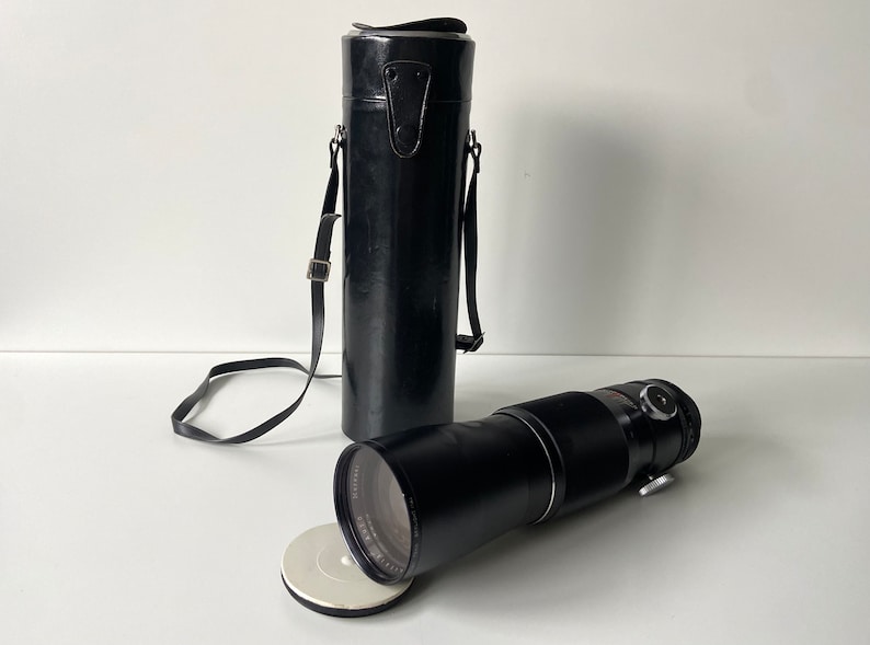 Puede incluir: Un objetivo de c&aacute;mara negro con una funda de transporte de cuero negro. El objetivo est&aacute; sobre una superficie blanca con una tapa circular blanca.