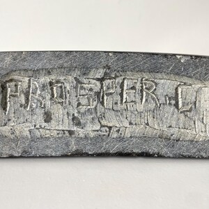 May include: A rectangular, gray stone object with the carved text "PAD SHER L". The stone has a rough, textured surface and appears to be a decorative piece or a small sculpture. The letters are deeply carved into the stone.