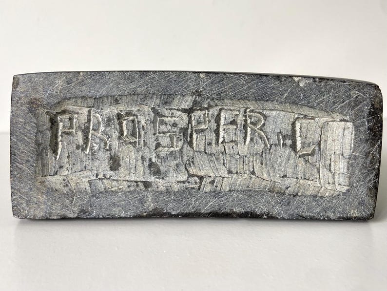 May include: A rectangular, gray stone object with the carved text "PAD SHER L". The stone has a rough, textured surface and appears to be a decorative piece or a small sculpture. The letters are deeply carved into the stone.