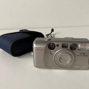 Puede incluir: Una cámara de película compacta Minolta Riva Zoom 115 de color plateado con sistema de lente Multi AF, flash integrado y botón de disparo en la parte superior. La cámara viene acompañada de un estuche protector azul oscuro con una correa negra.