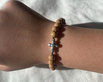 May include: A wooden bead bracelet with a silver cross charm. The bracelet features light brown, round wooden beads strung together with a silver cross pendant in the center. The bracelet is worn on a wrist.