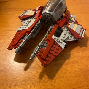 Op de afbeelding: Een rode en grijze Lego Star Wars V-4X-D-sterrenjager met een transparante cockpit.