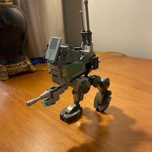 May include: A green and grey Lego model of an AT-RT walker with a black gun and two antennas. The walker is standing on a wooden surface.
