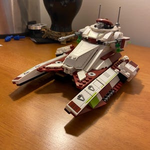 Puede incluir: Un caminante AT-TE de Lego Star Wars blanco y rojo con detalles verdes y una parte superior roja. El caminante tiene dos cañones en la parte delantera y dos en la parte trasera. El caminante está sentado sobre una superficie de madera.