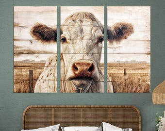 May include: A triptych wall art print depicting a close-up of a cow's face. The cow has a pale coat and brown ears. The background shows a field and a wooden fence. The artwork is mounted on wooden planks, creating a rustic aesthetic.