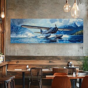 Può includere: Un dipinto di un idrovolante blu e bianco che vola sopra un lago con montagne sullo sfondo. Il dipinto è appeso a una parete in un ambiente da caffè con tavoli e sedie in legno.