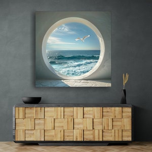 May include: A white circular window with a view of a blue ocean with white waves and a seagull flying in the sky. The window is set in a white wall with a stone floor.
