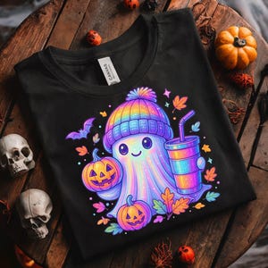 Könnte beinhalten: Schwarzes T-Shirt mit einem farbenfrohen Cartoon-Geist, der eine Regenbogenmütze trägt und Kürbisse und ein Getränk hält. Herbstblätter und eine Fledermaus ergänzen das Halloween-Thema.