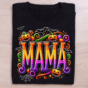 Puede incluir: Camiseta negra con un dise&ntilde;o de Halloween. La palabra "MAMA" se muestra en letras grandes y coloridas, rodeada de calabazas, murci&eacute;lagos y telara&ntilde;as. El dise&ntilde;o utiliza acentos naranjas, morados y verdes, creando un aspecto festivo.