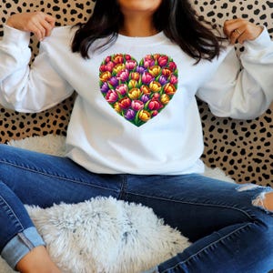 Pode incluir: Camisola branca com um desenho em forma de cora&ccedil;&atilde;o preenchido com tulipas coloridas. As tulipas s&atilde;o em tons de vermelho, rosa, amarelo, roxo e verde. A camisola &eacute; de manga comprida e tem gola redonda.